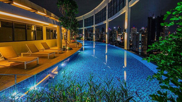 Une piscine moderne située sur le toit, avec des chaises longues et une vue sur les toits de la ville illuminée la nuit.