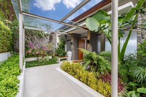 A modern outdoor area with a glass walkway, stone walls, and lush greenery including various plants and flowers. - Villa Narissa (Kamala)