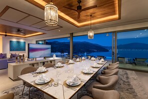 A modern dining room with a long table set for a meal, large windows offering a view of the mountains, and a television on the wall. - Villa Narissa (Kamala)