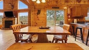 Interior de una cabaña de madera con una gran mesa de comedor, bancos de madera, una chimenea de piedra y un televisor.