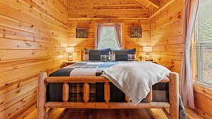 Un dormitorio de cabaña de madera con una cama, dos apliques de pared, un ventanal con cortinas y un adorno en la pared.