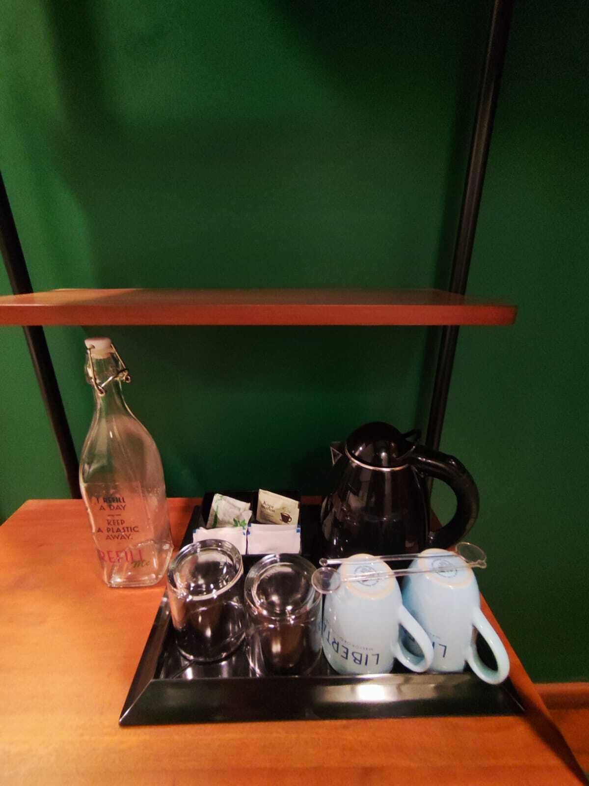 Un plateau avec une bouteille en verre, une bouilloire noire, deux tasses et une théière, le tout posé sur une table en bois devant un mur vert.