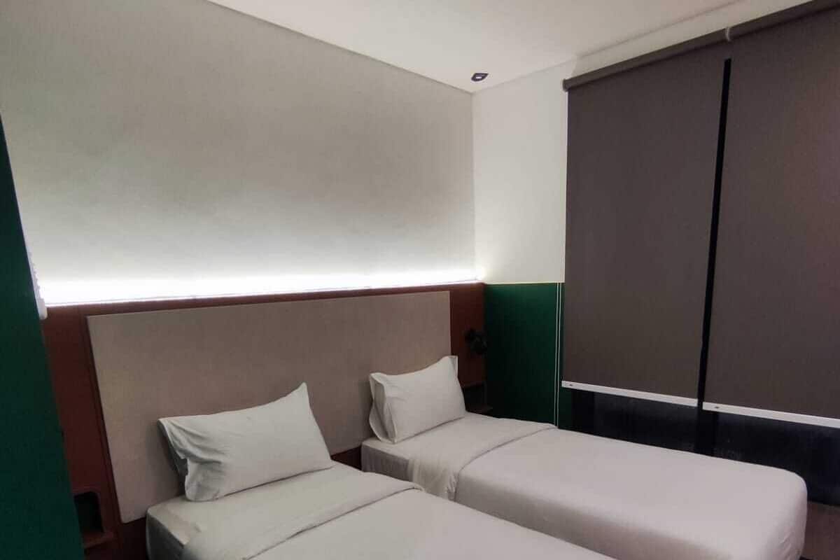 A hotel room with two single beds, a large window with blinds, a dark-colored wardrobe, and a green accent wall.