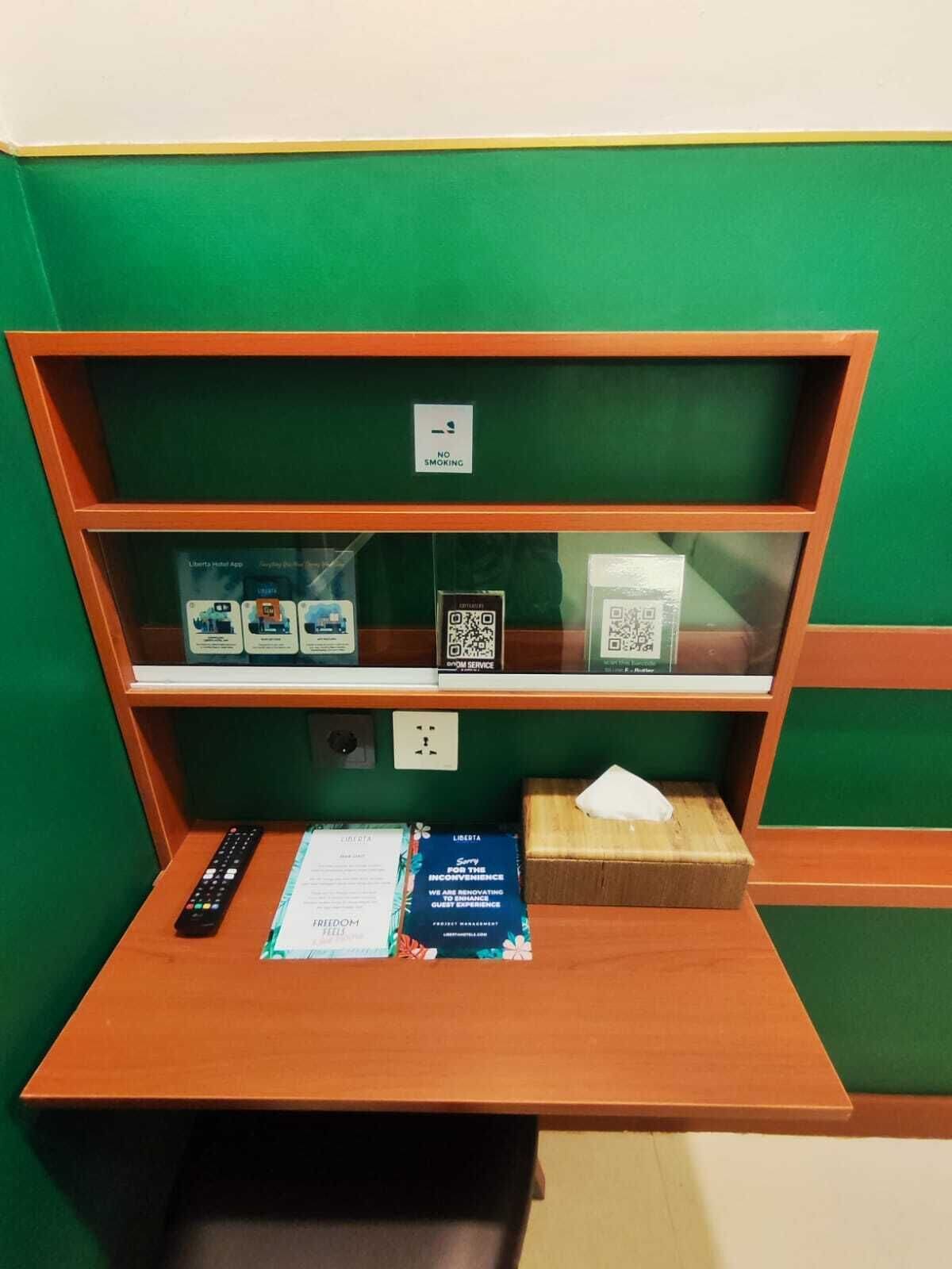 Habitación de hotel con pared verde, un escritorio de madera, un control remoto, una caja de pañuelos, un pañuelo, un folleto y un cartel de 'No Fumar'.