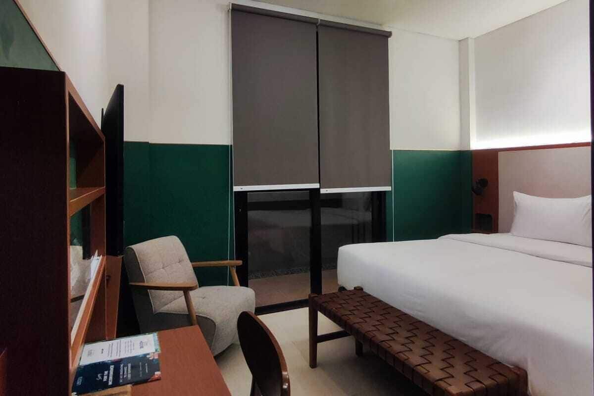 A hotel room with a bed, a desk with a chair, a bookshelf, and a window with blinds.