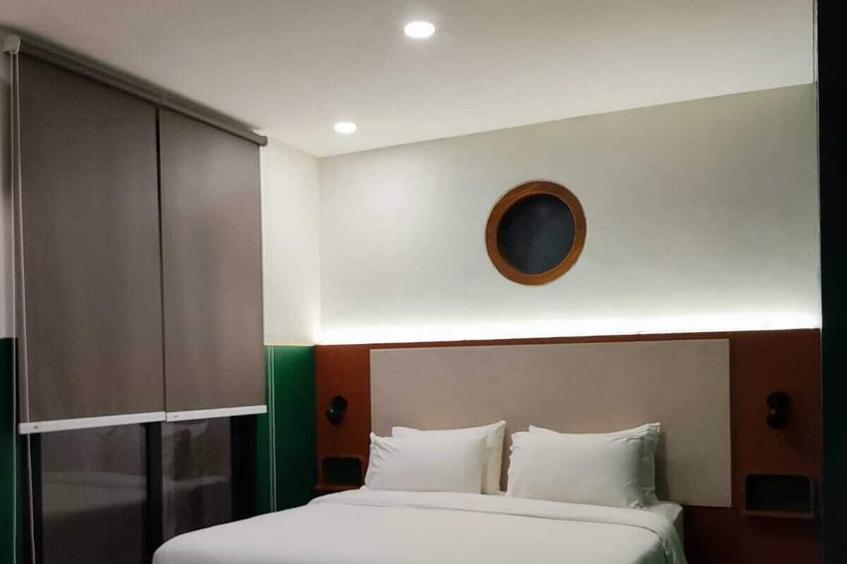 A modern hotel room with a large bed, a small desk, and a window with blinds.