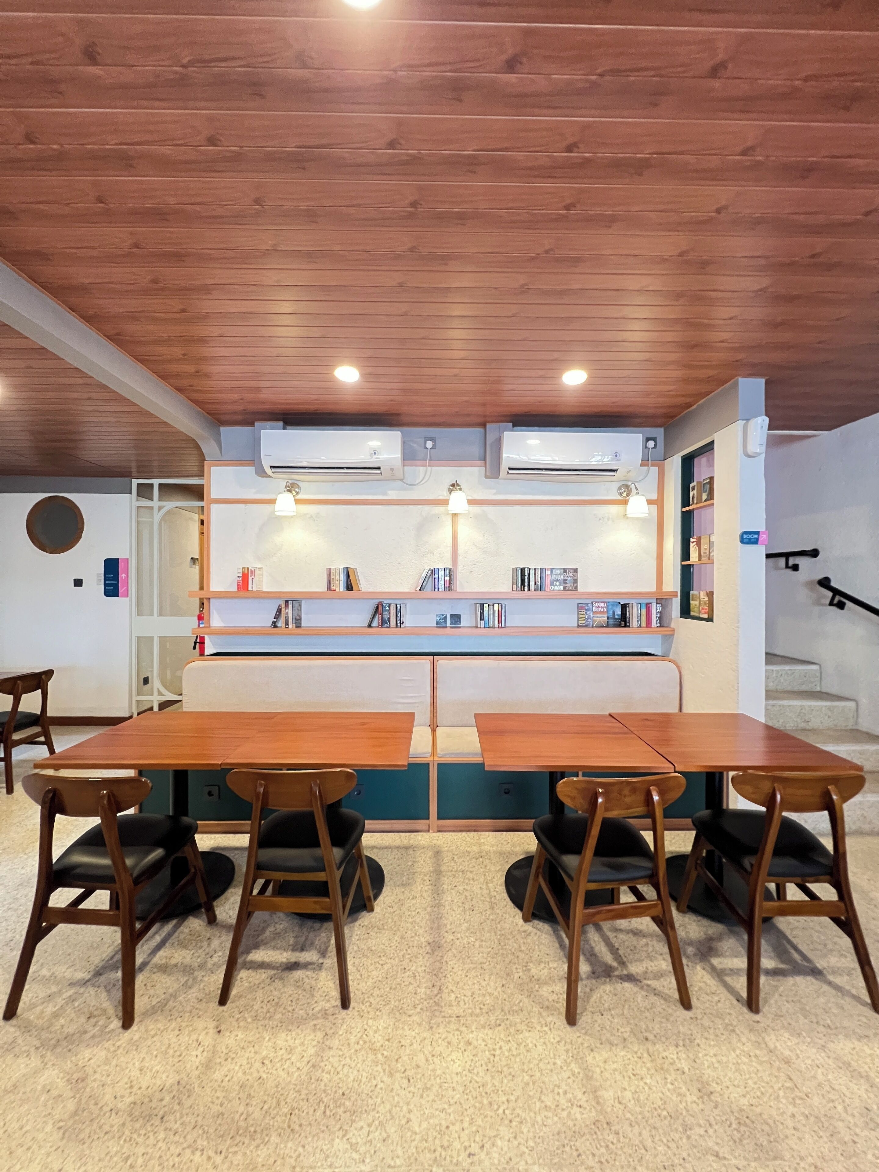 A cozy room with a wooden ceiling, two air conditioning units, a bookshelf, and a seating area with a table and chairs.