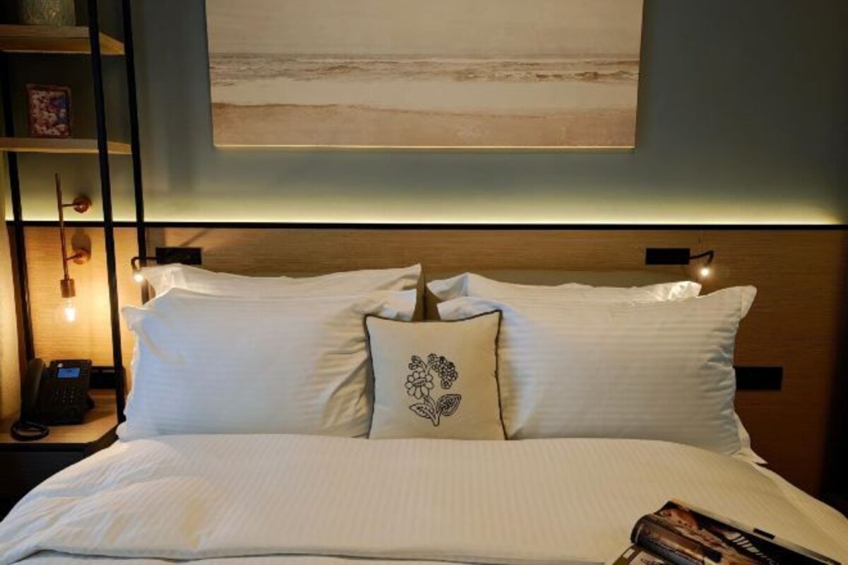 A neatly made bed with white linens and a decorative pillow, a bedside phone, and a wall-mounted artwork depicting a beach scene.