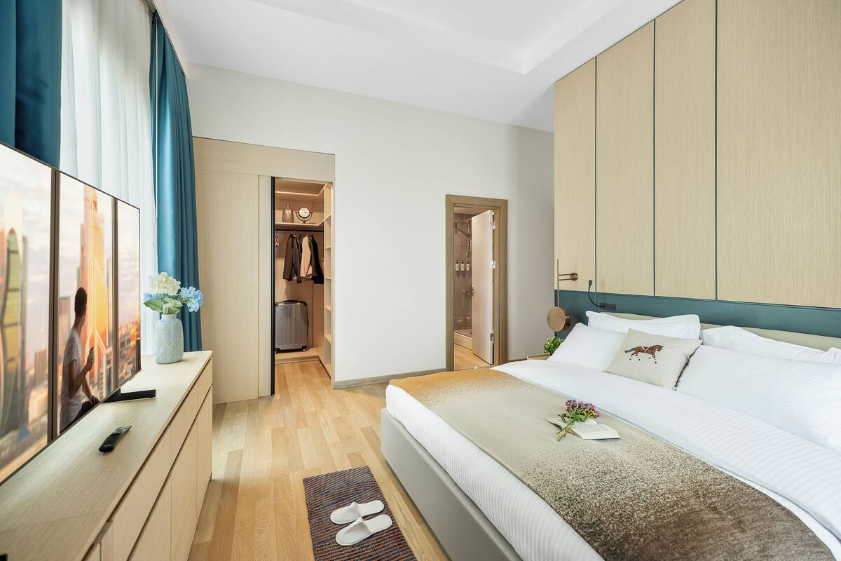 Ein modernes Hotelzimmer mit einem großen Bett, einem begehbaren Kleiderschrank und Blick auf die Stadt durch große Fenster.