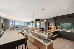 Een moderne hotel eetruimte met een lange gedeelde tafel, een buffet en uitzicht op de skyline door grote ramen.