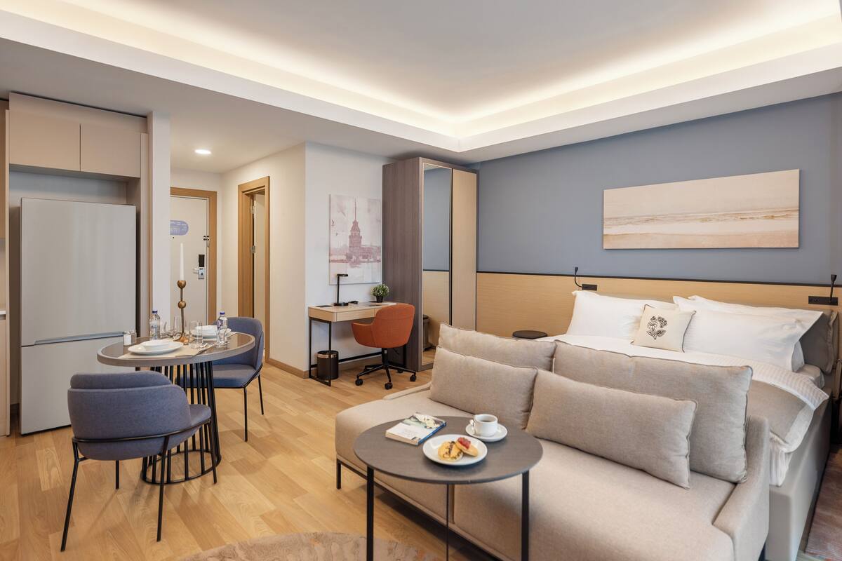 Ein modernes Hotelzimmer mit Bett, Sofa, Esstisch mit Stühlen, Schreibtisch und einer kleinen Küchenzeile.
