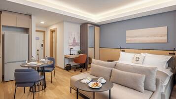 A modern hotel room with a bed, a sofa, a dining table with chairs, a desk, and a small kitchen area.