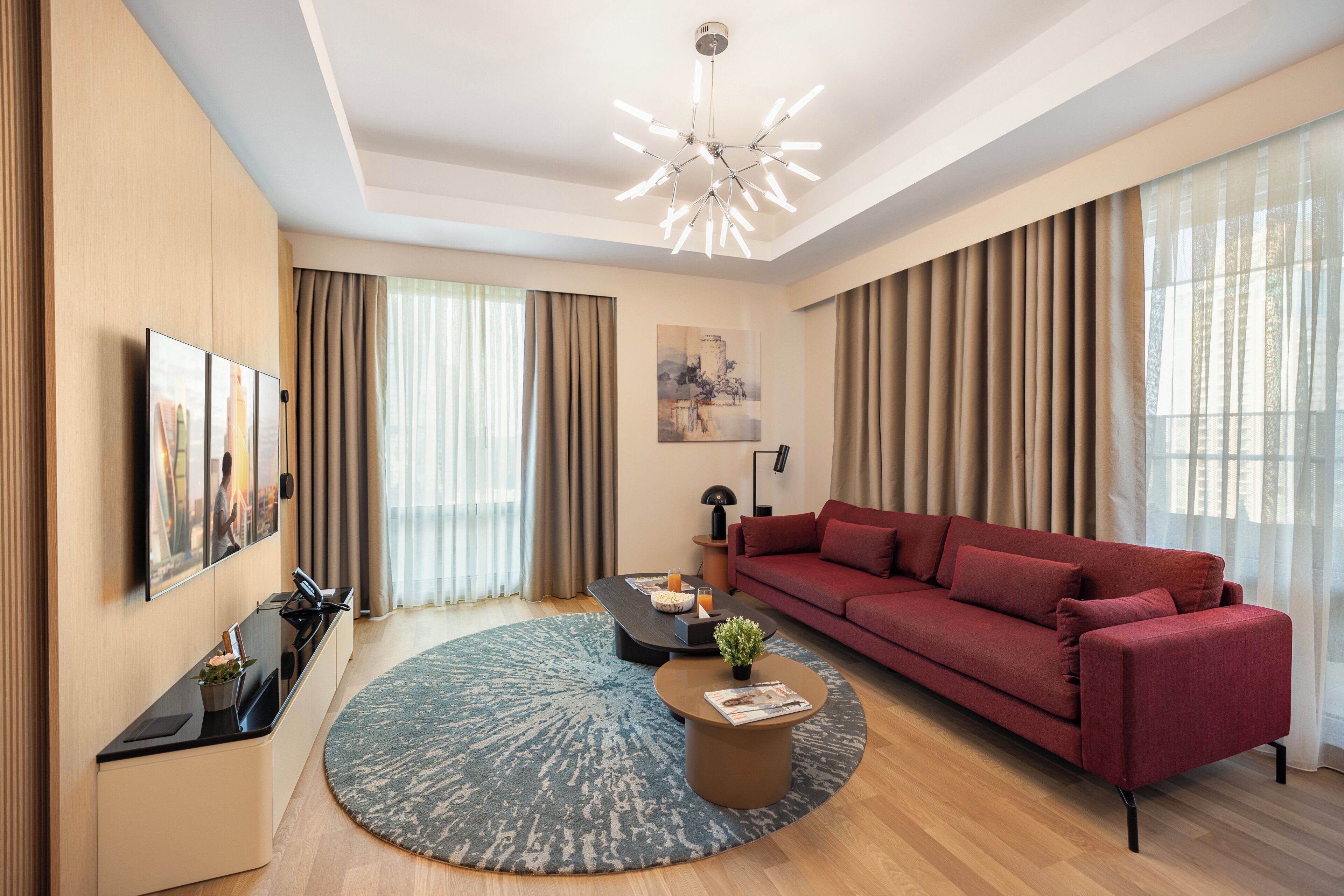 A modern living room with a red sofa, a round coffee table, a flat-screen TV, and a large window with curtains.
