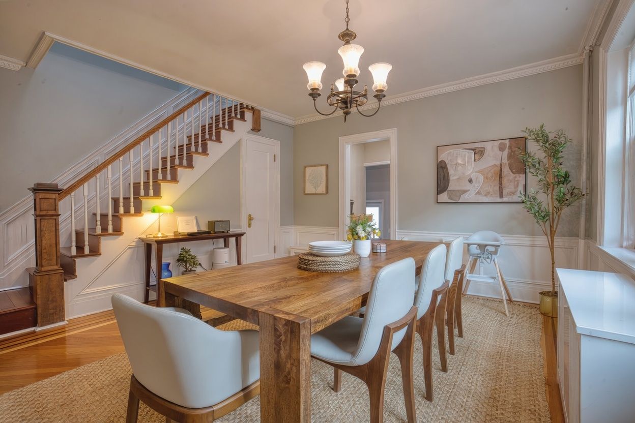 A dining area with a wooden table, white chairs, a chandelier, and a staircase.