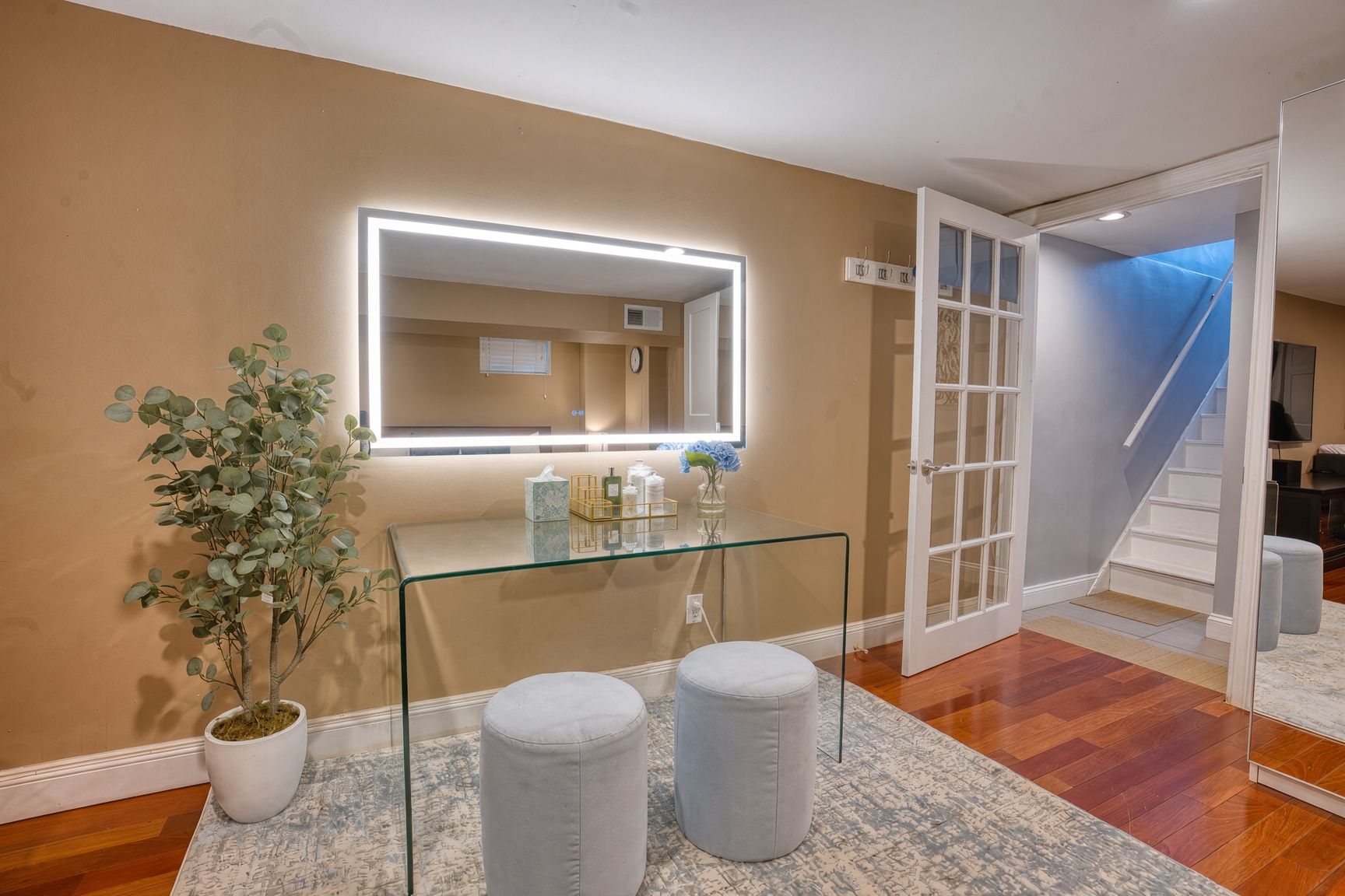 A hallway with a glass table, two ottomans, a potted plant, a mirror with lighting, and a staircase.
