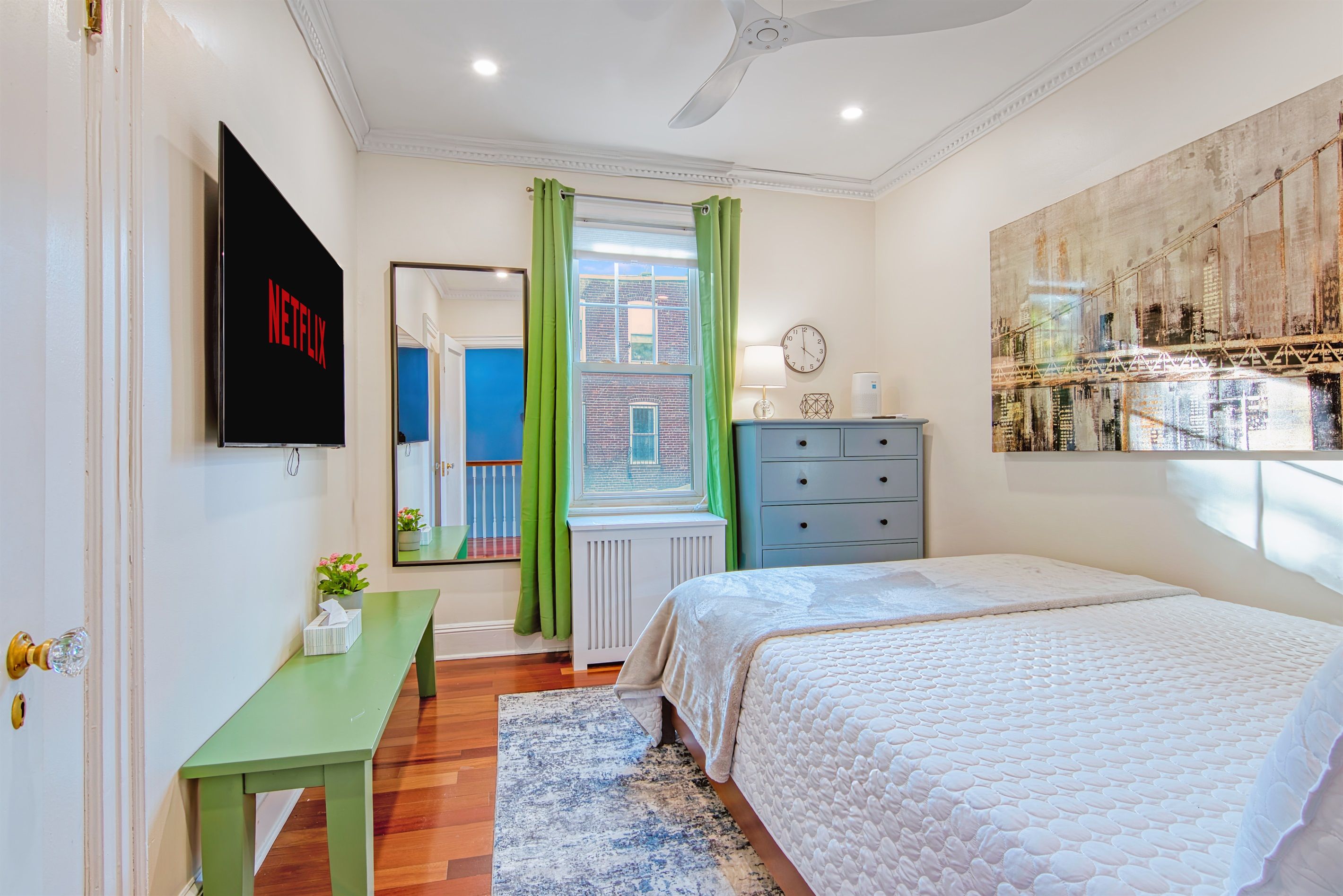 A bedroom with a bed, a dresser, a TV mounted on the wall, a window with green curtains, and a radiator.