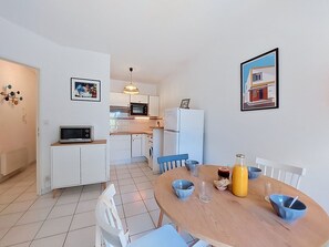 A compact kitchen with white cabinets, a microwave, and a refrigerator. There is a dining table with chairs and a small TV on a cabinet. - Parc de Pontaillac by Interhome (Vaux Sur Mer)