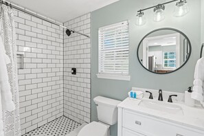 A bathroom with white subway tiles, a shower with a glass door, a toilet, a sink with a mirror, and a window with blinds.