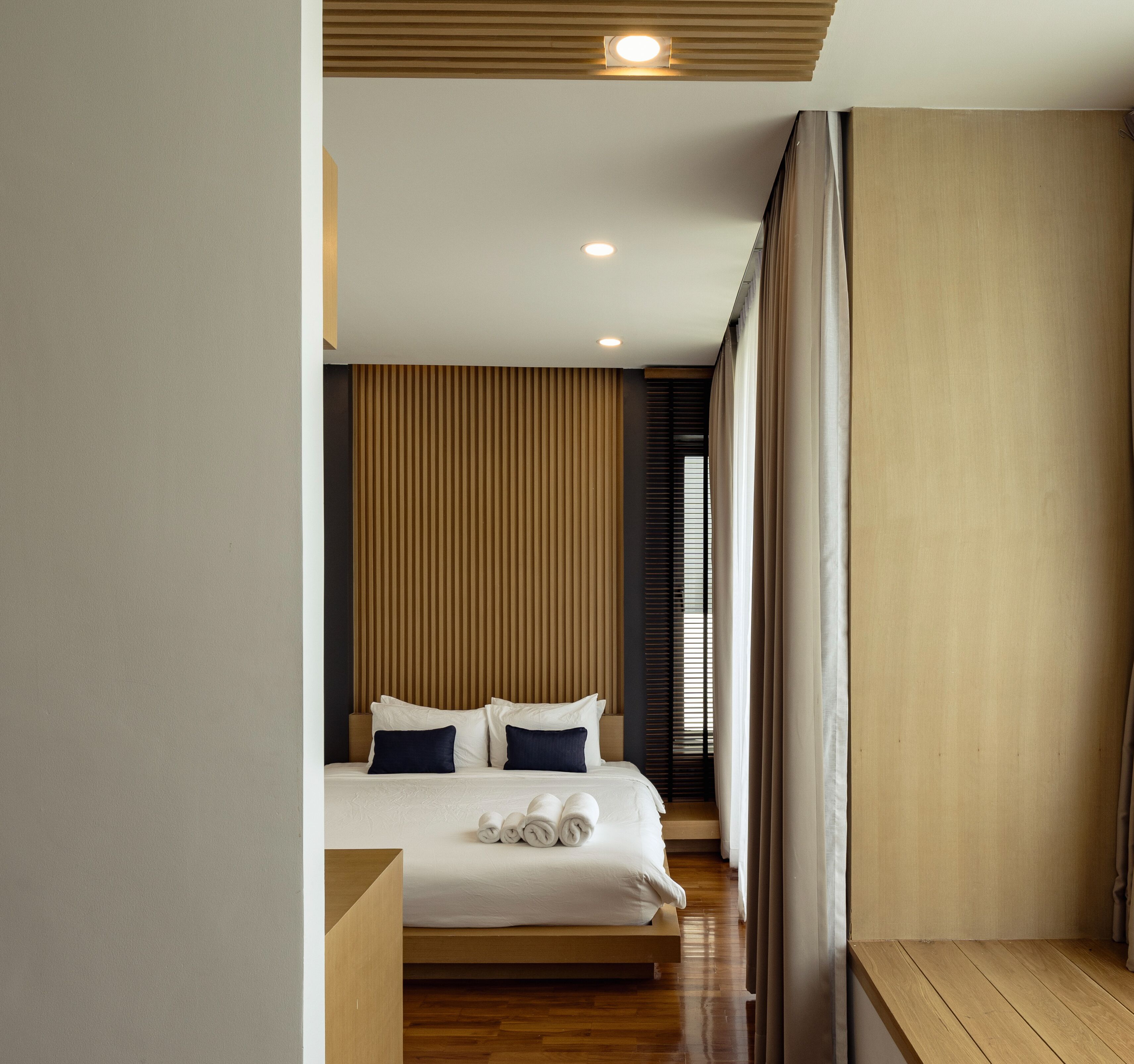 Una habitación de hotel moderna con suelo de madera, una cama con sábanas blancas y almohadas oscuras, cabecero de madera y un ventanal con persianas.