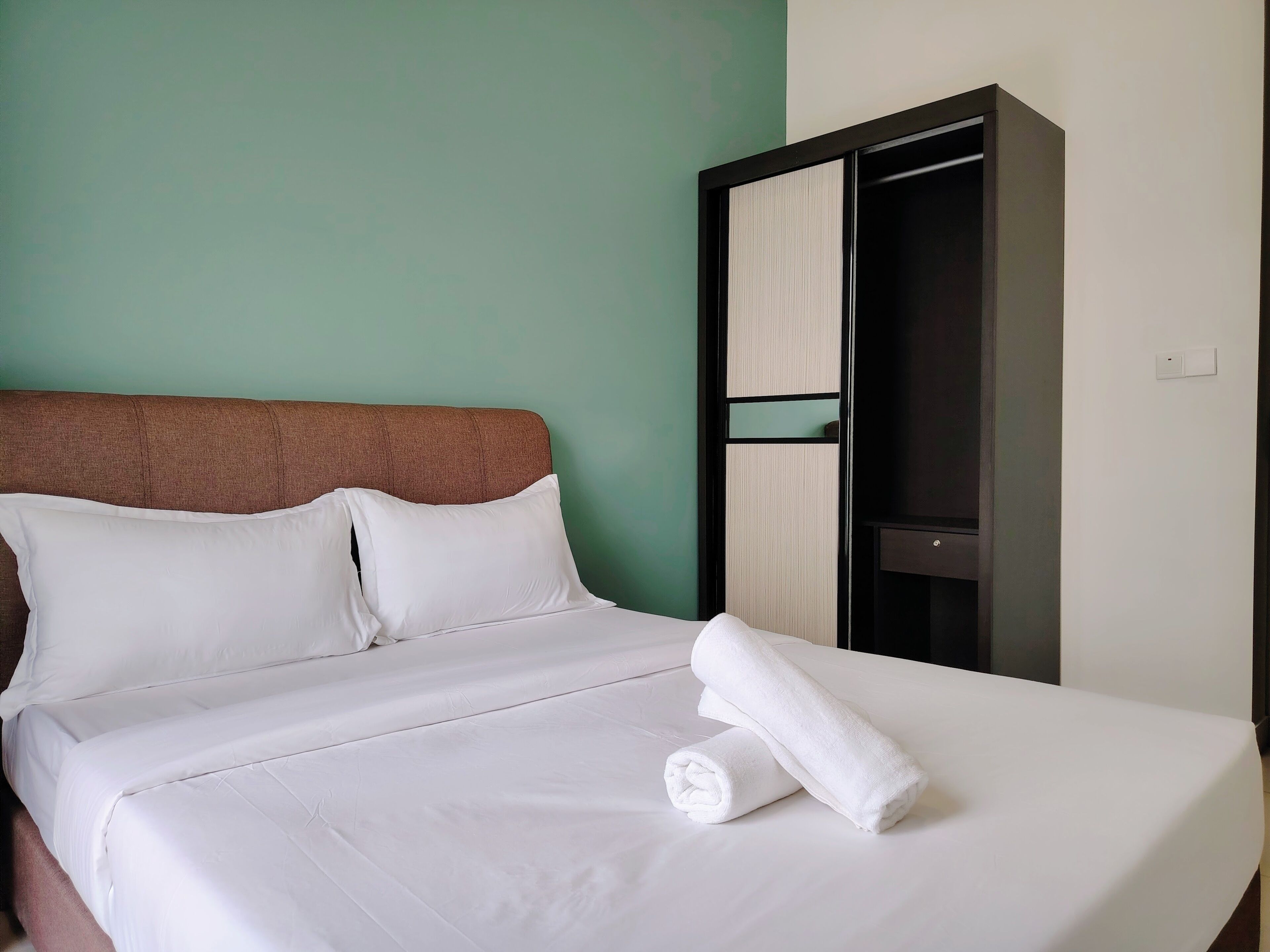 Una cama bien hecha con sábanas y almohadas blancas, una toalla blanca enrollada y un armario de madera oscura con espejo.