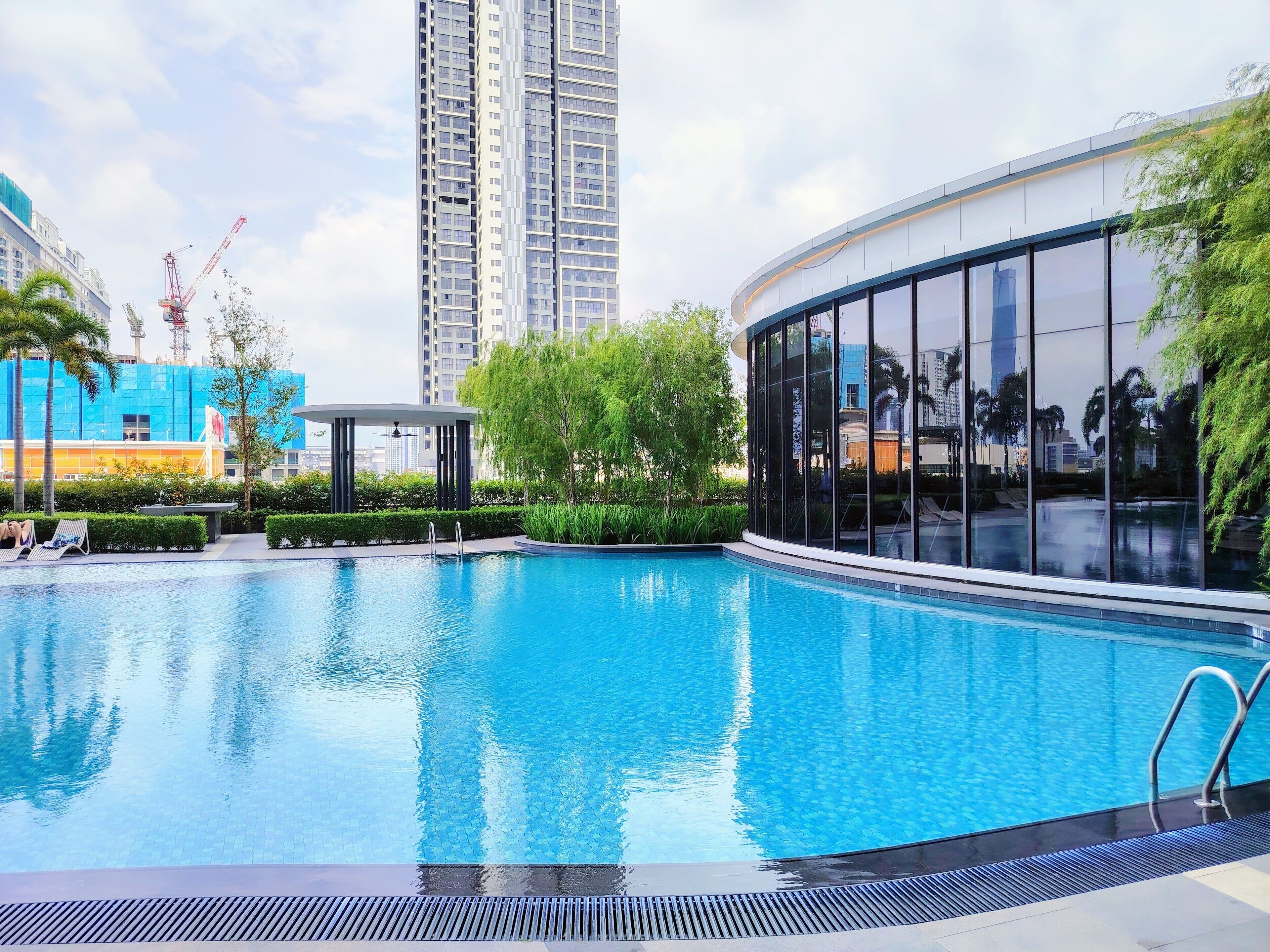 A swimming pool with a tiled deck, surrounded by greenery and modern buildings.