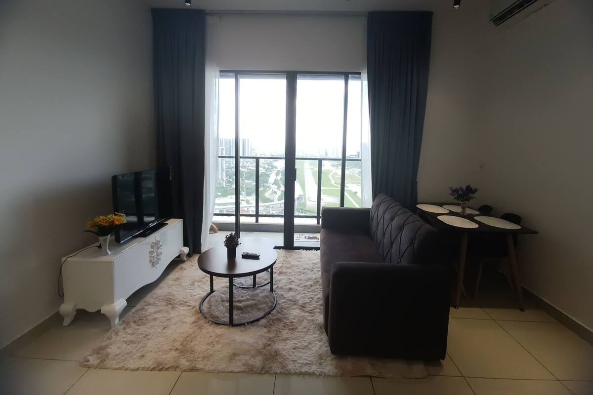A living room with a sofa, a TV, a small table, and a balcony with a view of the city.