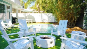 A backyard with a swimming pool, white Adirondack chairs, a fire pit, and a white fence.