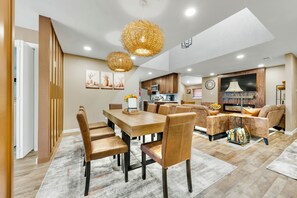 A spacious dining area with a wooden table and chairs, a kitchen in the background, and a living area with a sofa and a TV. - A&M Cabin-Pool Table Deck BBQ-10mins Kyle Field (Bryan)