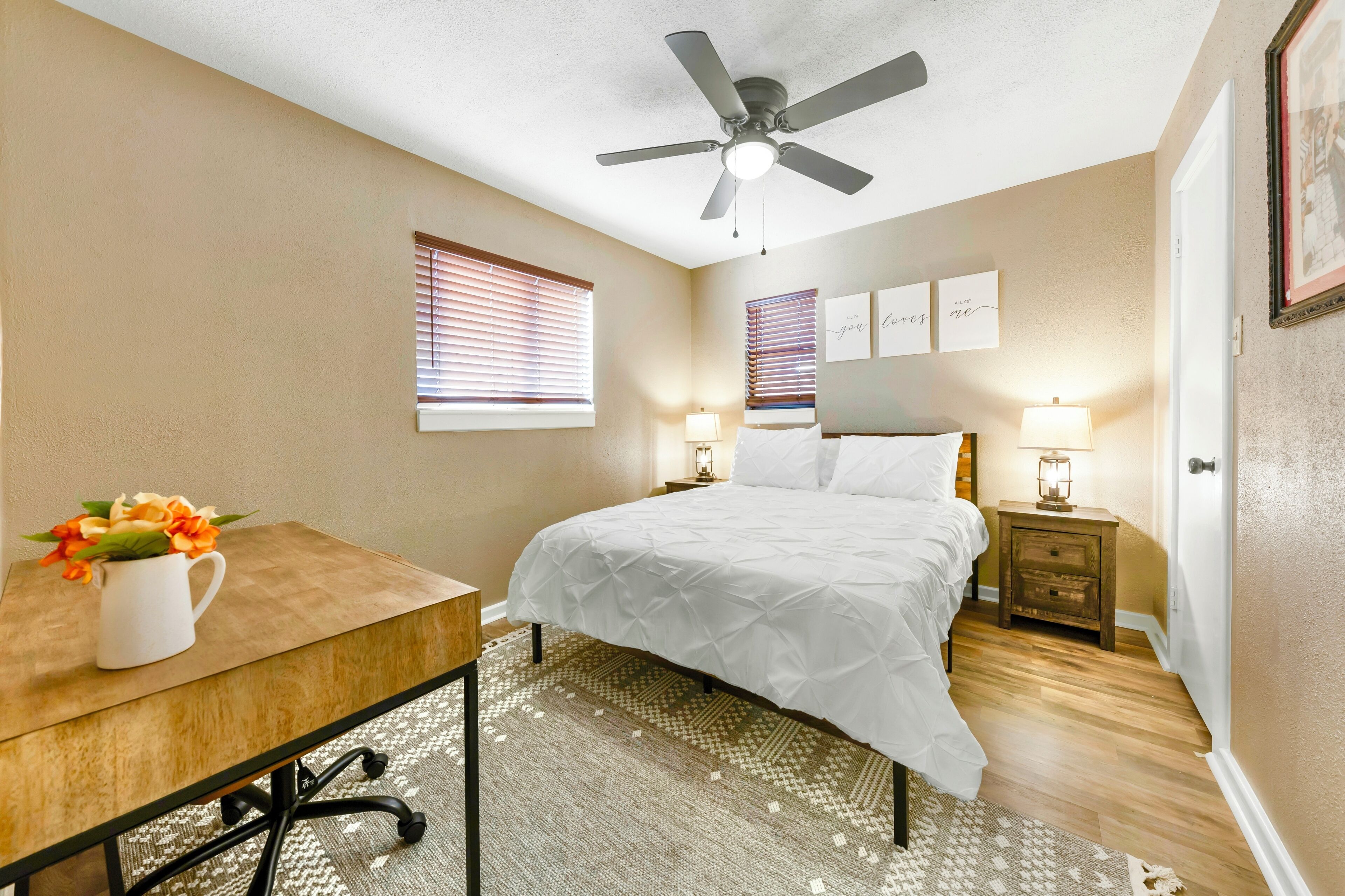 A bedroom with a bed, a desk, a ceiling fan, a nightstand with a lamp, and a window with blinds.