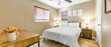 A bedroom with a bed, a desk, a ceiling fan, a nightstand with a lamp, and a window with blinds.