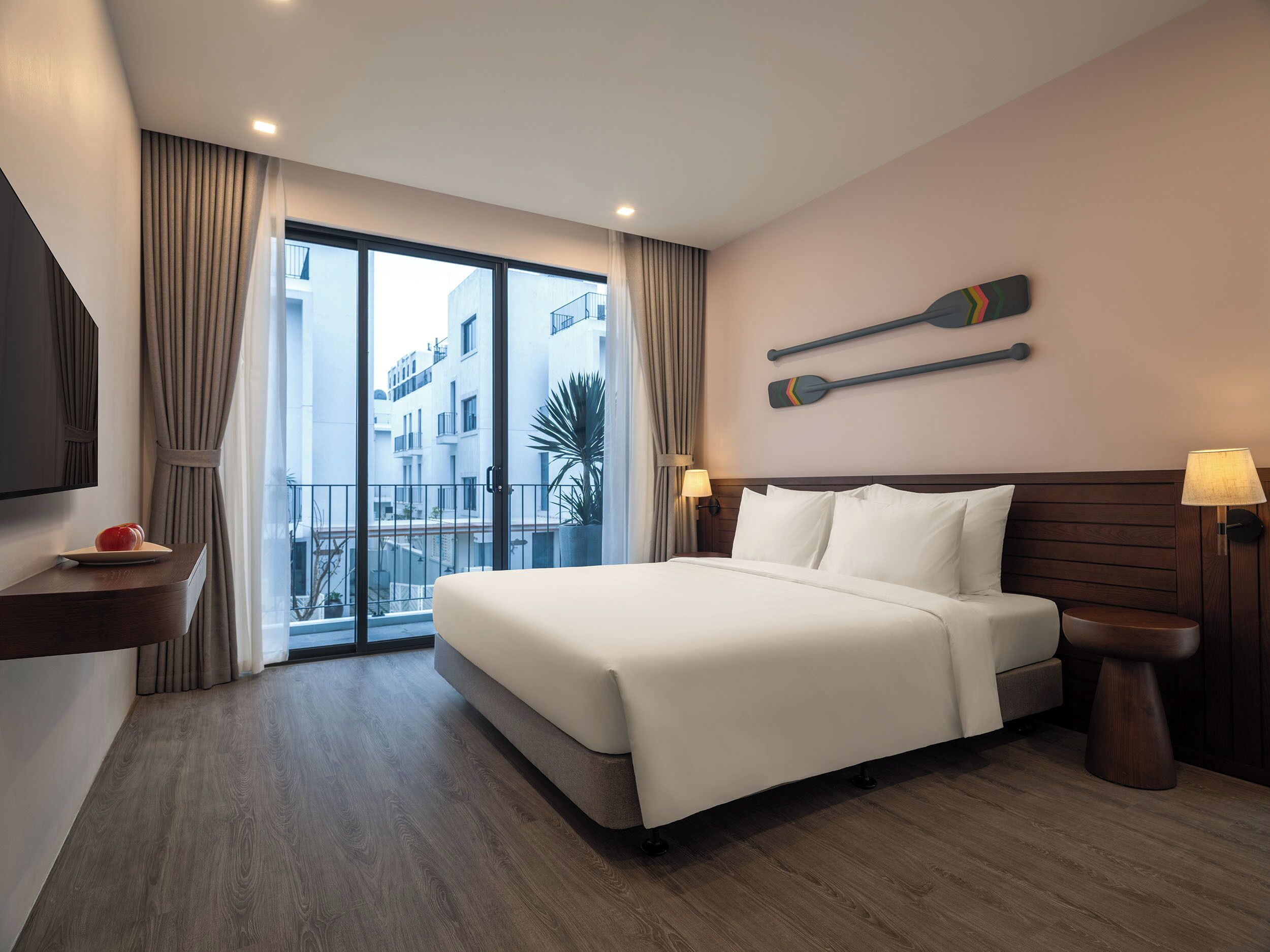 A hotel room with a large bed, a TV, a wooden floor, and a balcony with curtains.