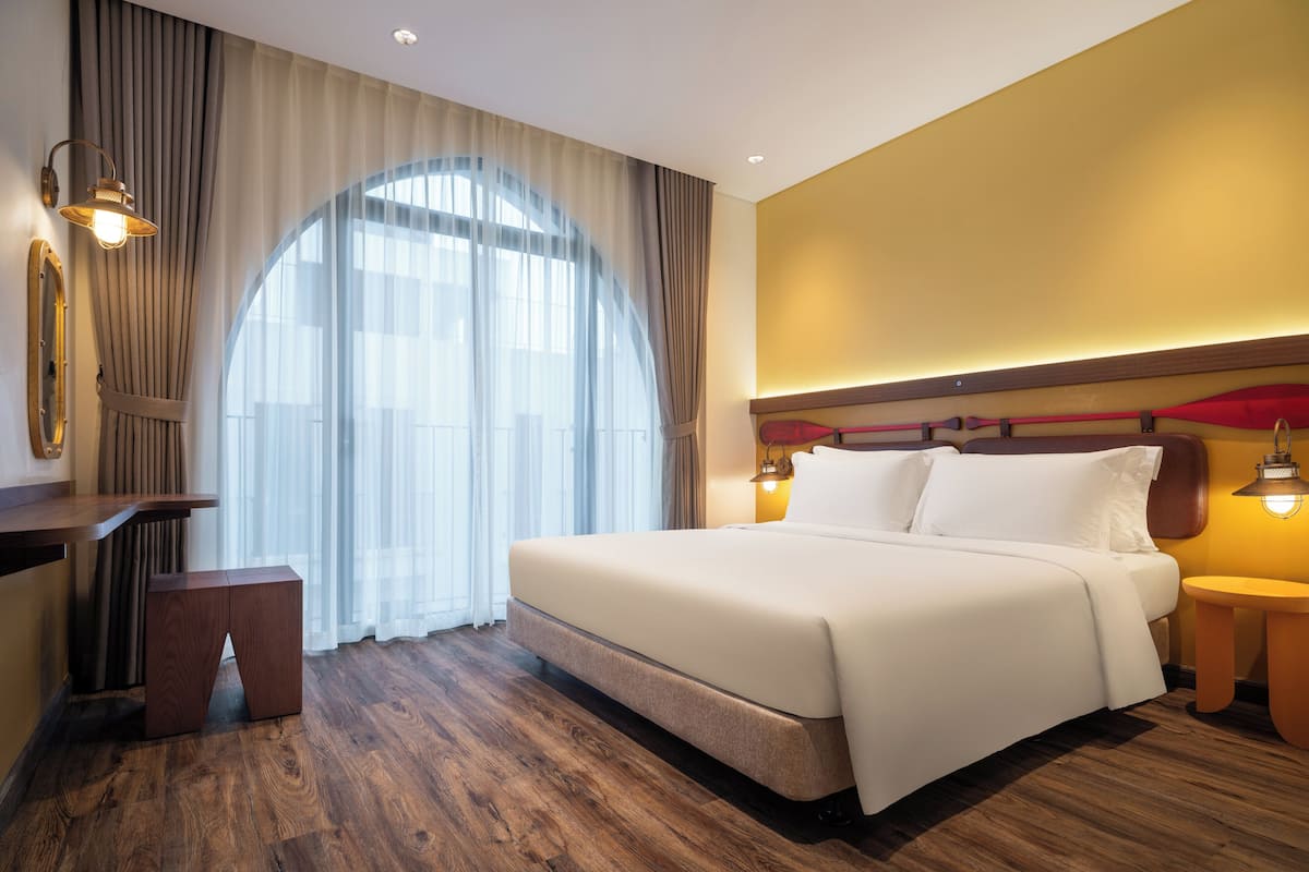 A hotel room with a large bed, a desk, and a chair. There is a window with curtains and a view of the outdoors.