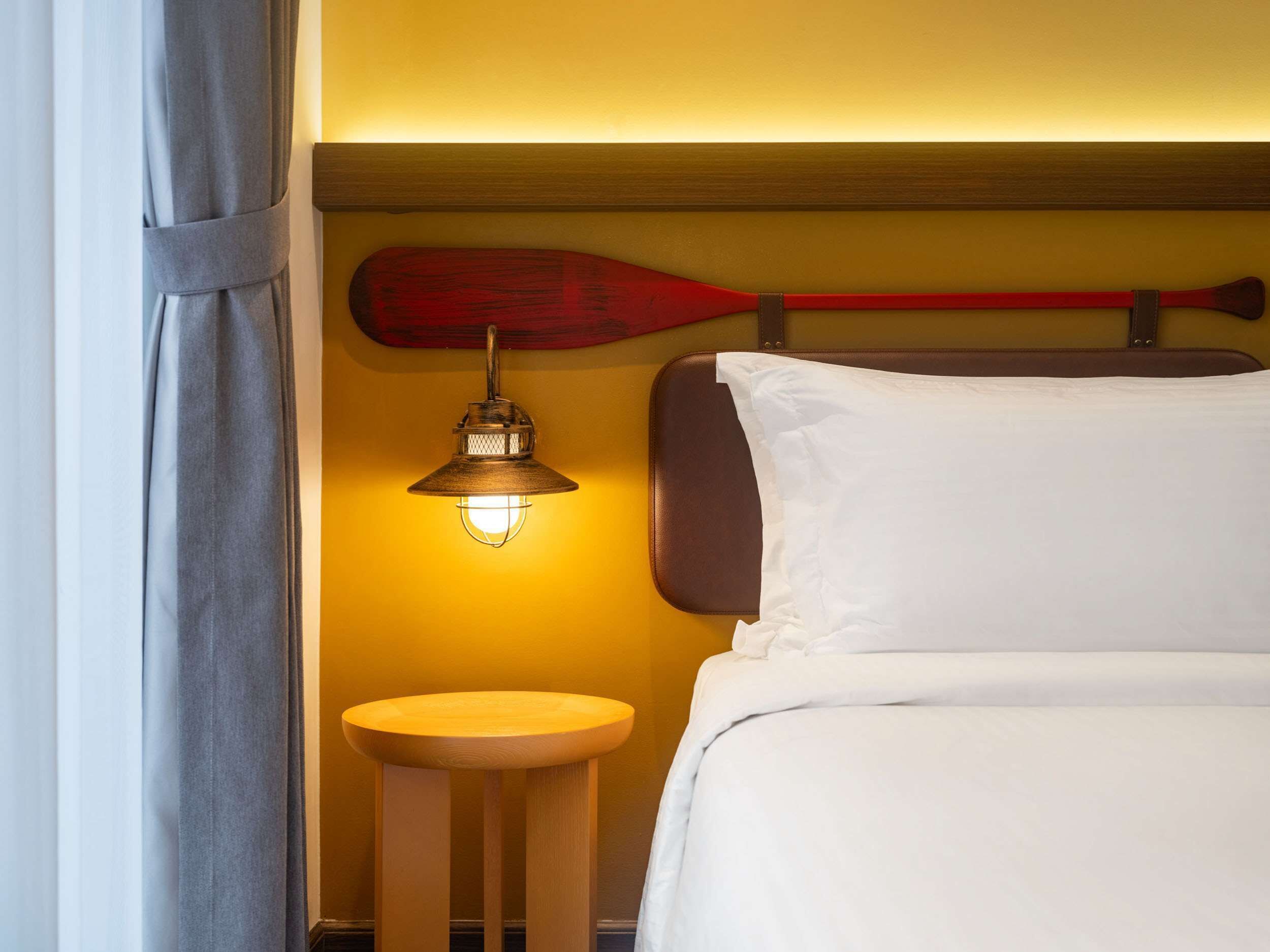A hotel room with a bed, a small wooden stool, a wall-mounted light fixture, and a red paddle leaning against the wall.
