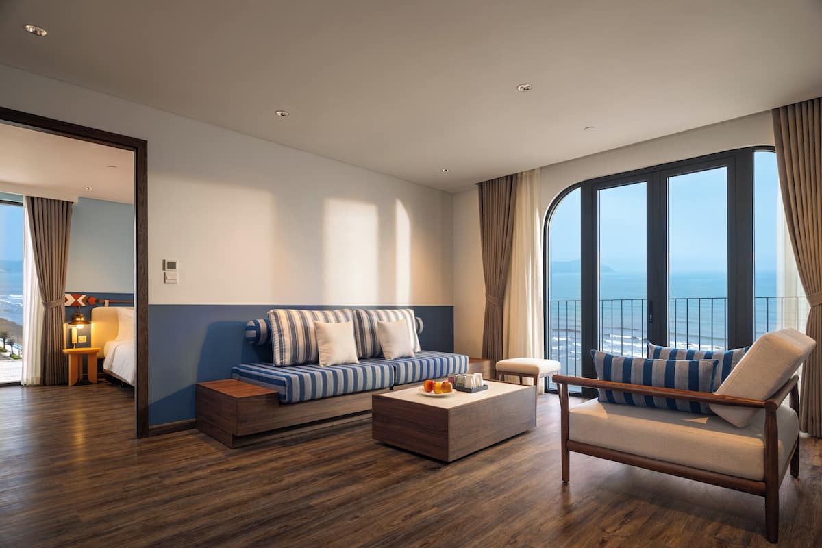 A modern hotel room with a wooden floor, a striped sofa, a cushioned armchair, a small table with fruits, and a large window offering a view of the ocean.