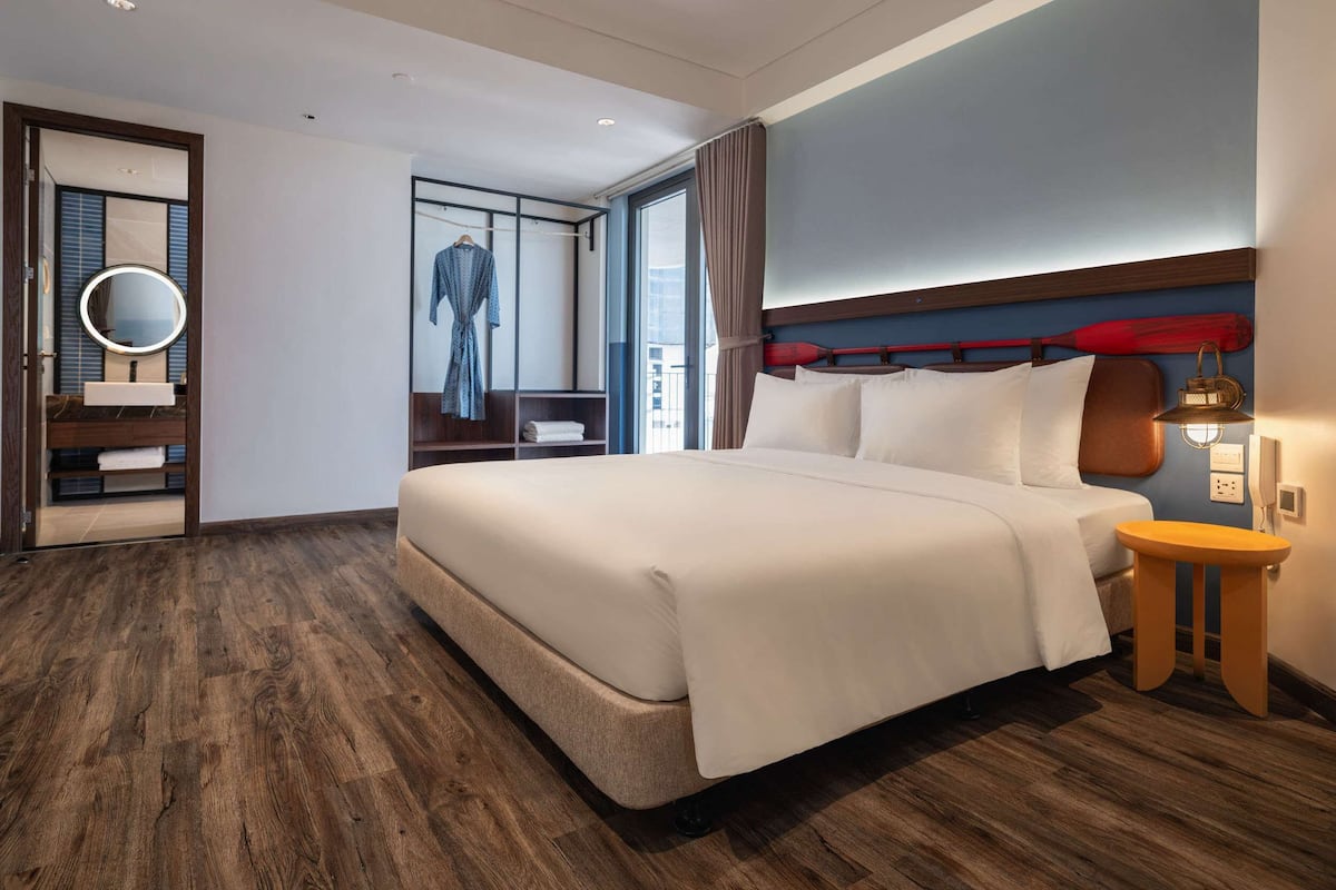 A modern hotel room with a large bed, a wooden floor, a bathroom with a mirror, and a view of the city.