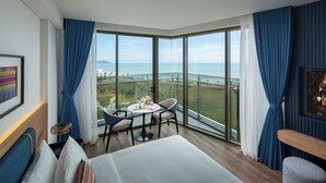 A hotel room with a large window, a bed, a small table with chairs, and a view of the sea. - Lasong Hotel & Villas Sam Son By The Unlimited Collection (Nam Sam Son)