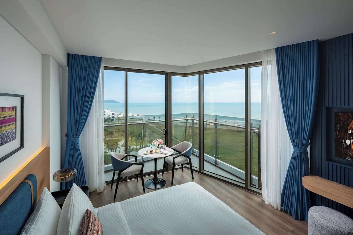 A hotel room with a large window, a bed, a small table with chairs, and a view of the sea.