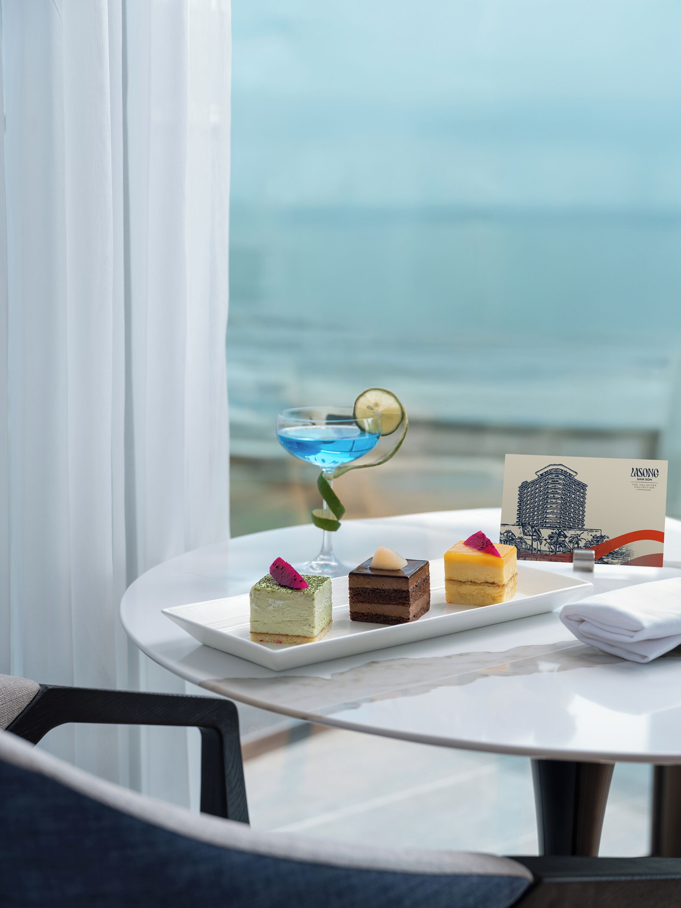 A table with desserts and a cocktail, a folded napkin, and a business card, with a view of the sea in the background.