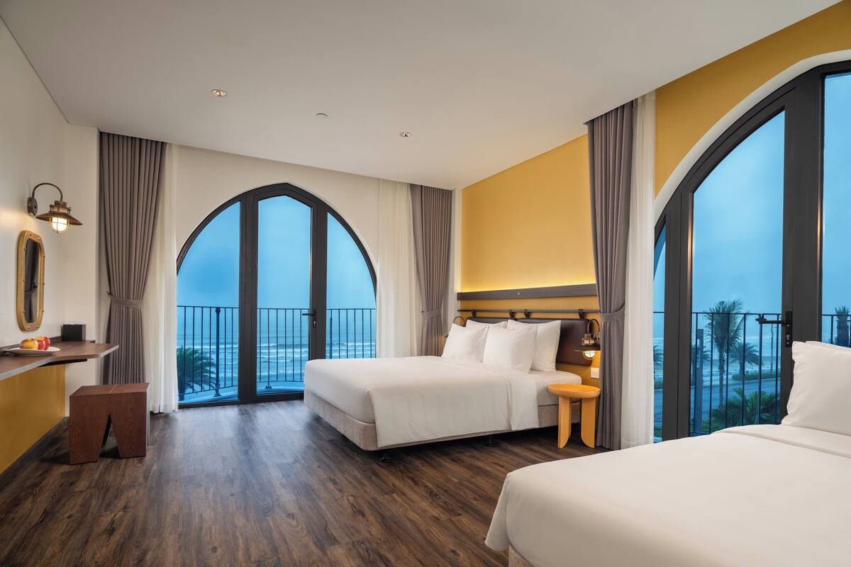 A hotel room with two beds, a desk, and a large arched window offering a view of the ocean.