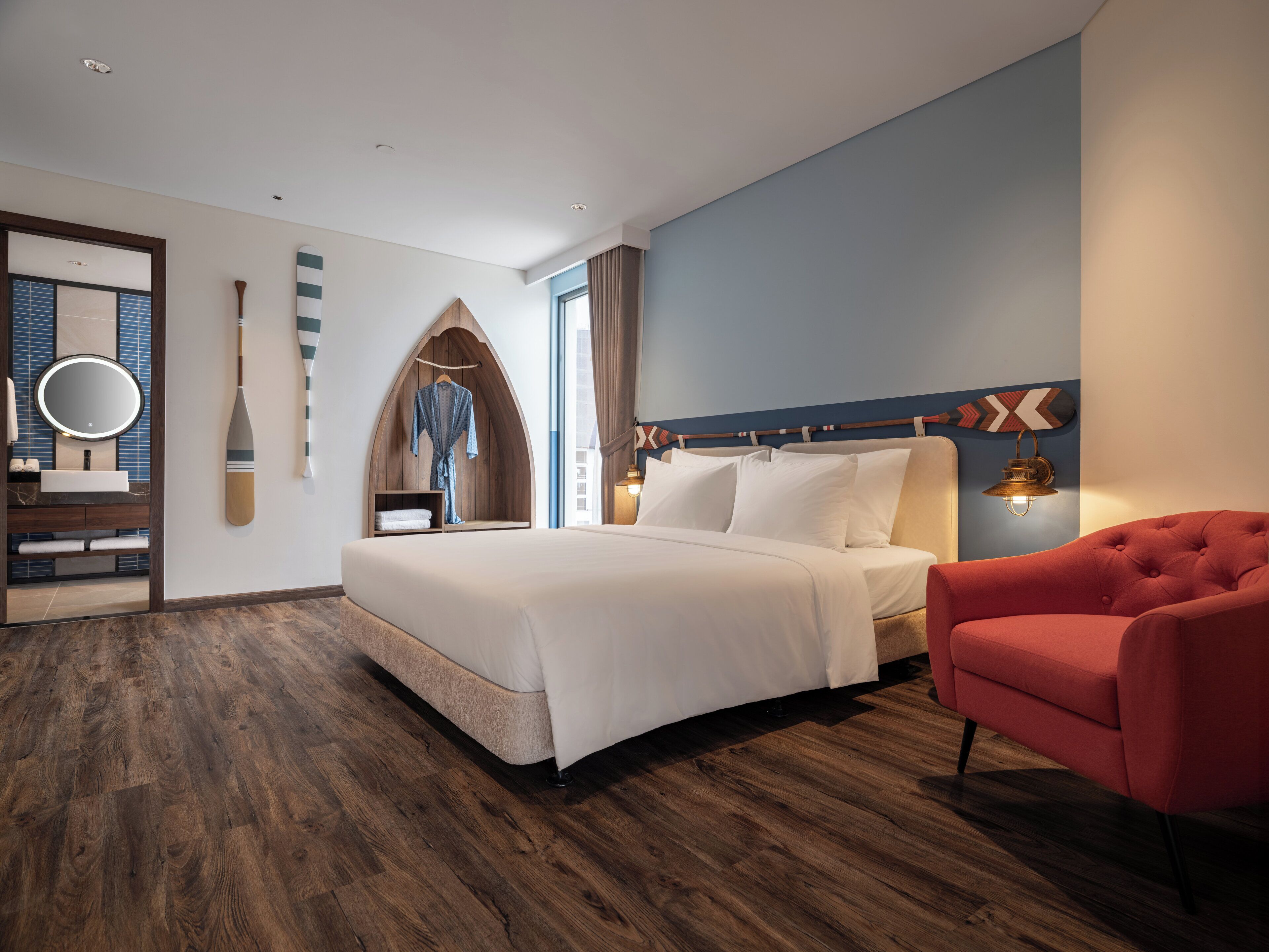 A modern hotel room with a large bed, a red armchair, a wooden arched wall, and a large window.
