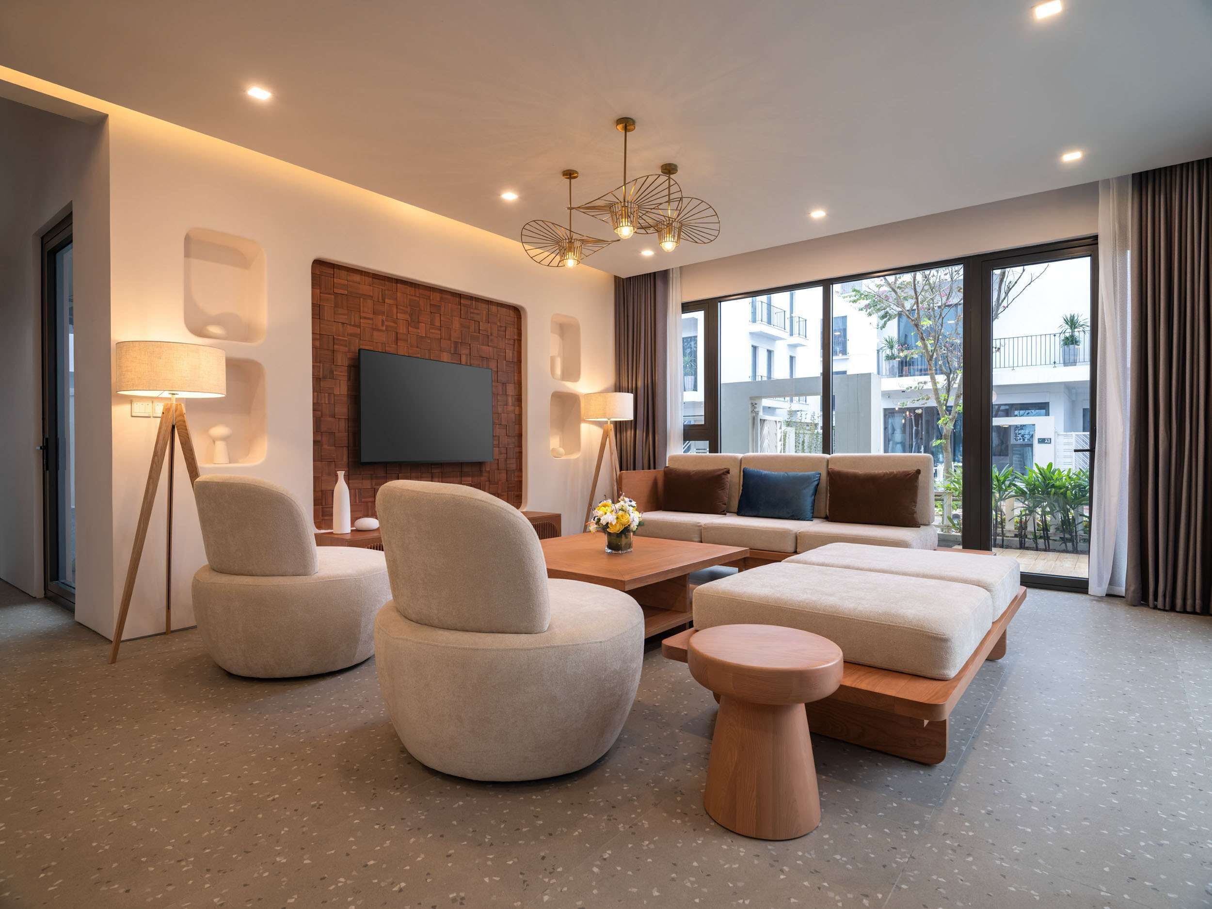Ein modernes Wohnzimmer mit einer Sofagarnitur, einem Couchtisch und zwei Sesseln. An der Wand ist ein Flachbildfernseher angebracht, außerdem steht eine Stehlampe und eine Pendelleuchte im Raum. Große Fenster bieten einen Ausblick nach außen.