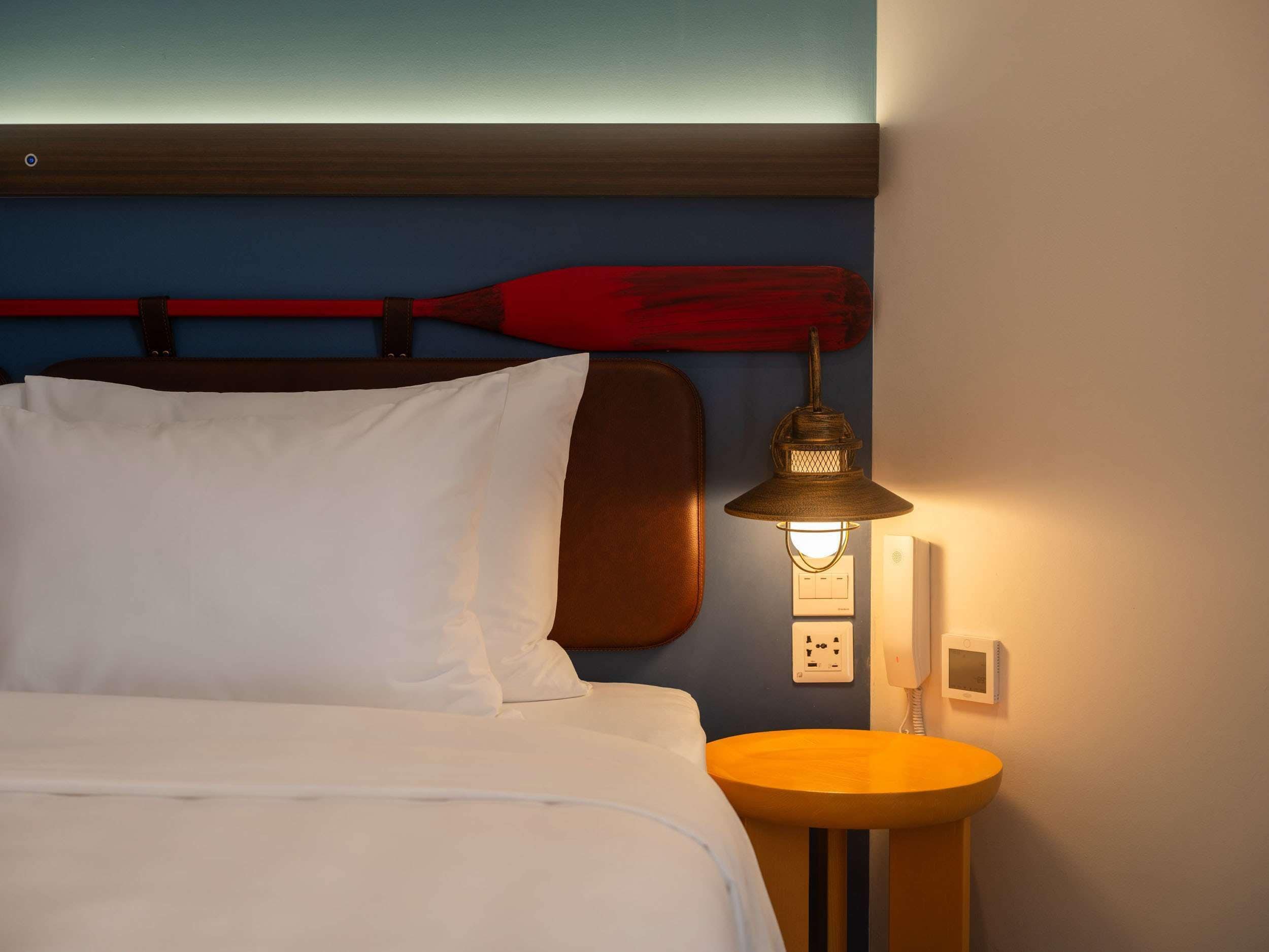 A neatly made bed with white linens, a red paddle leaning against the wall, a wall-mounted light fixture, and a small round table with a yellow surface.