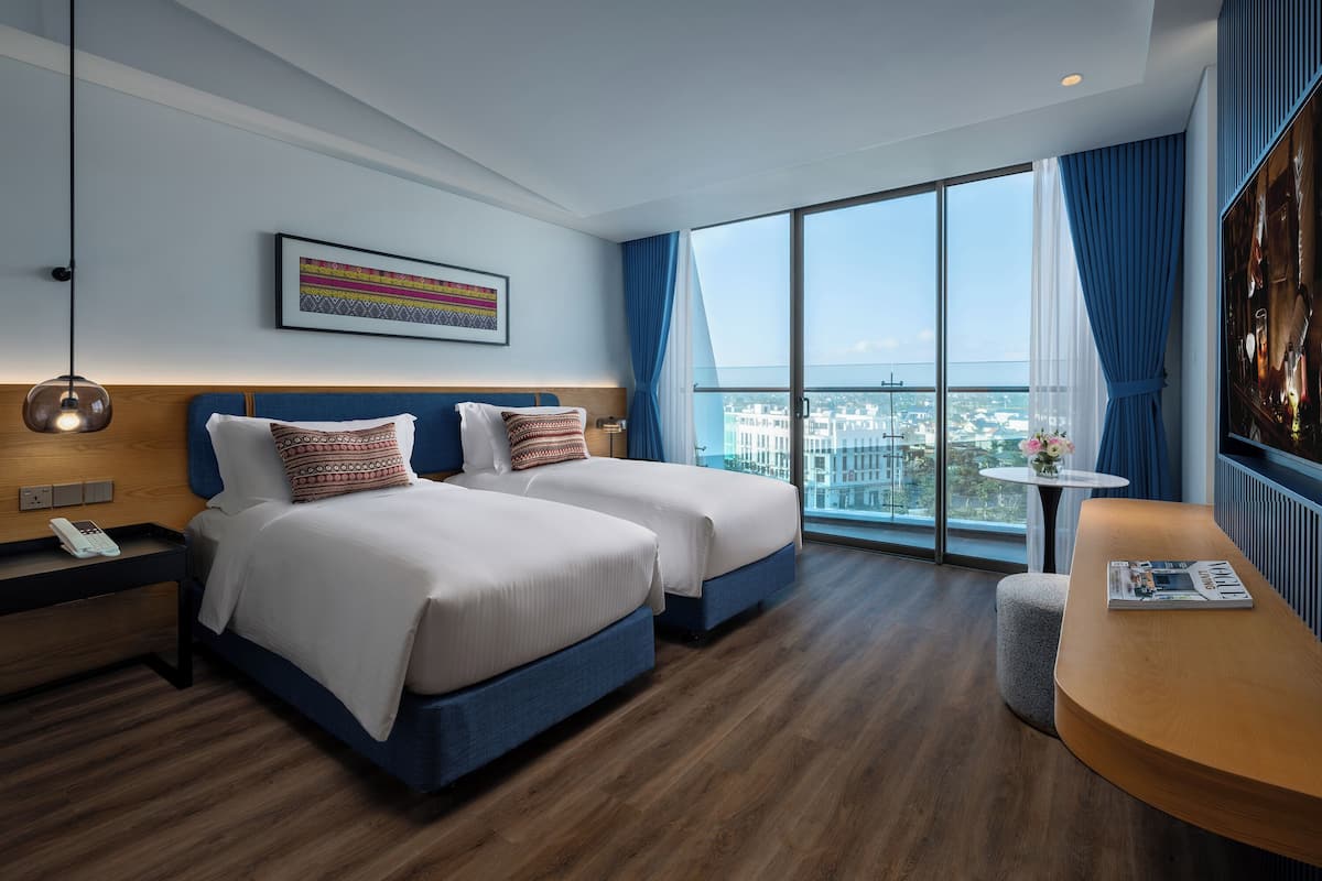A hotel room with two beds, a large window with a city view, a small table with a magazine, and a wall-mounted phone.