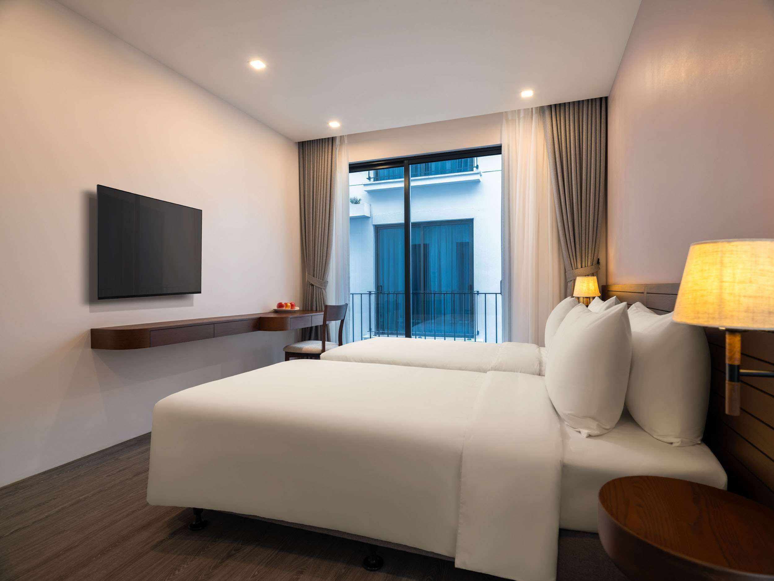 A hotel room with a bed, a TV, a bedside table, a lamp, and a view of the outside balcony.
