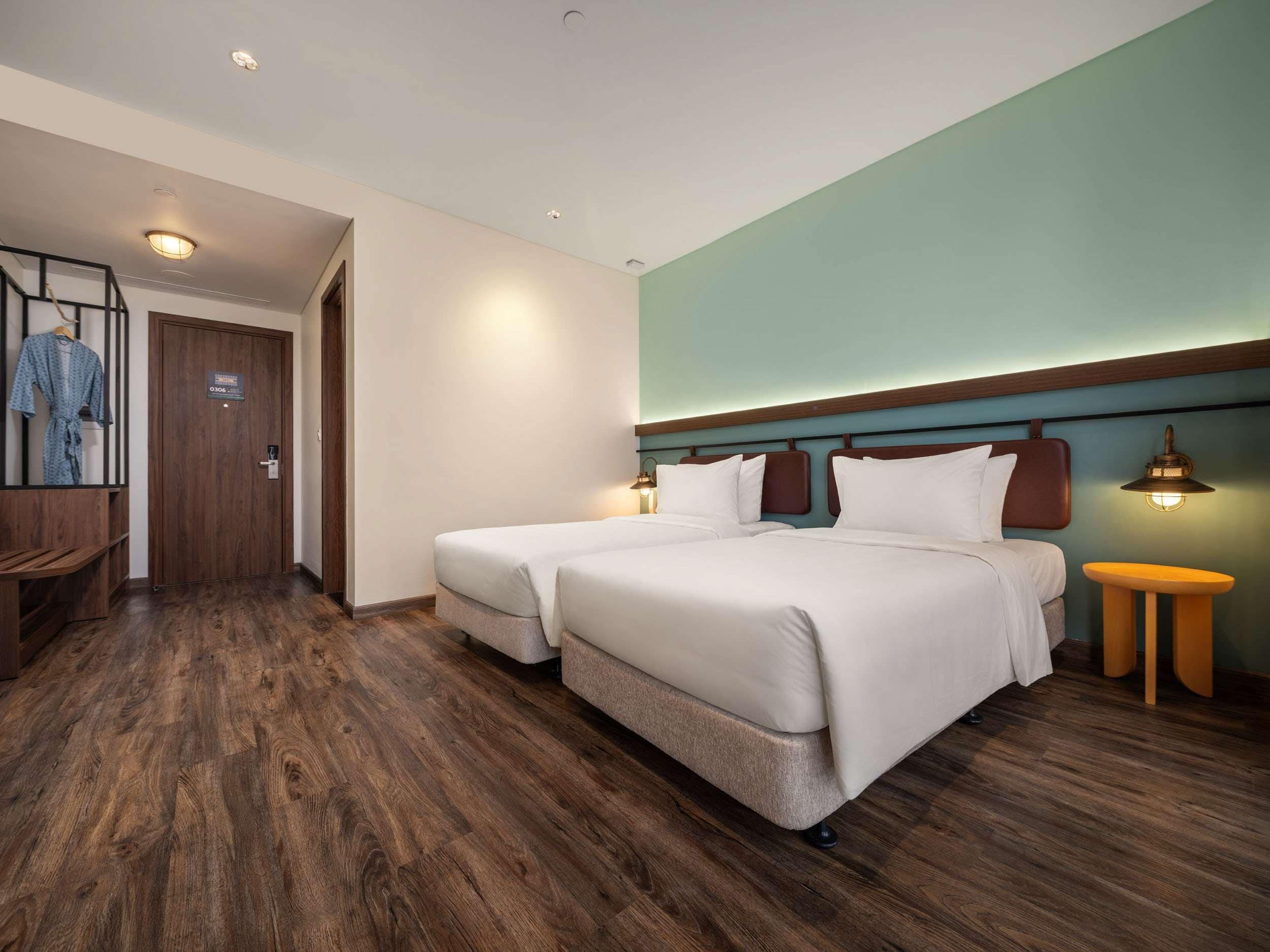A hotel room with two beds, a wooden floor, a green accent wall, and a small table with a lamp.