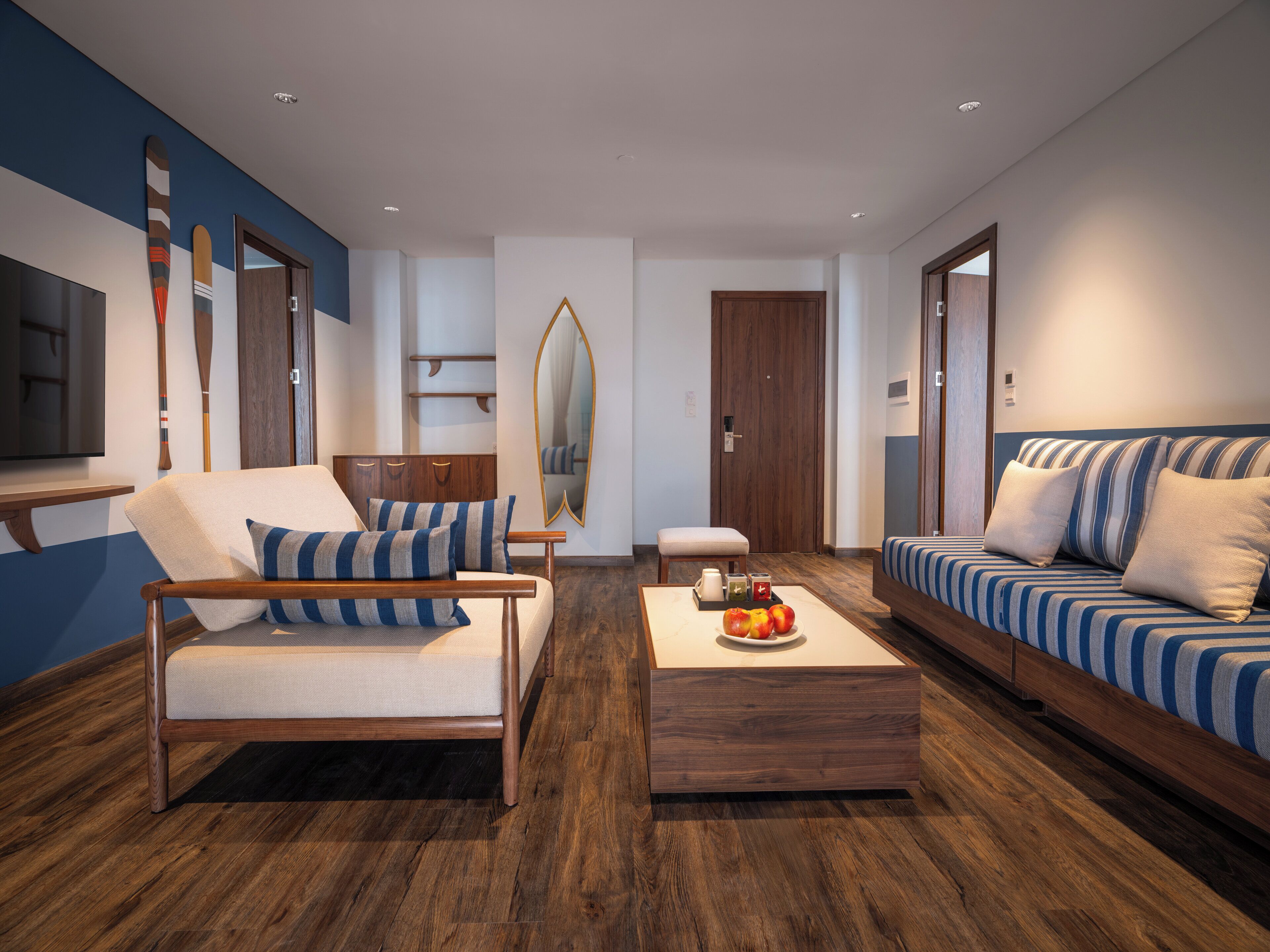 A modern living room with a wooden floor, a sofa with striped cushions, a chair with striped cushions, a coffee table with fruit, and a large mirror.