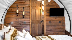 Una habitación tipo tienda acogedora con paredes de madera, una cama con ropa de cama blanca, dos lámparas fijadas a la pared y un televisor en la pared.