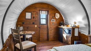 A cozy, circular tent-style room with wooden walls, a dining table with chairs, a small kitchenette, and a bed with a white blanket.