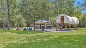 Un campamento con carromatos cubiertos, una mesa de picnic y sillas, una pequeña construcción con techo y una zona de grava.