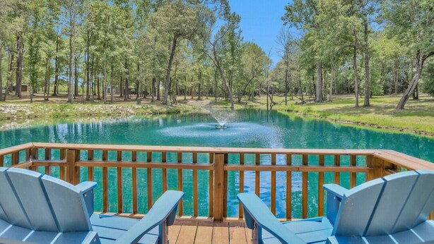 Una terraza de madera con sillas azules da a un estanque azul y cristalino, rodeado de árboles.